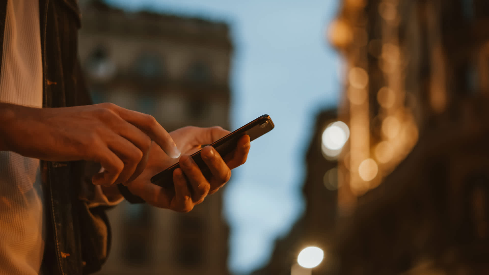 Person using smartphone in city at dusk with blurred urban lights in background.