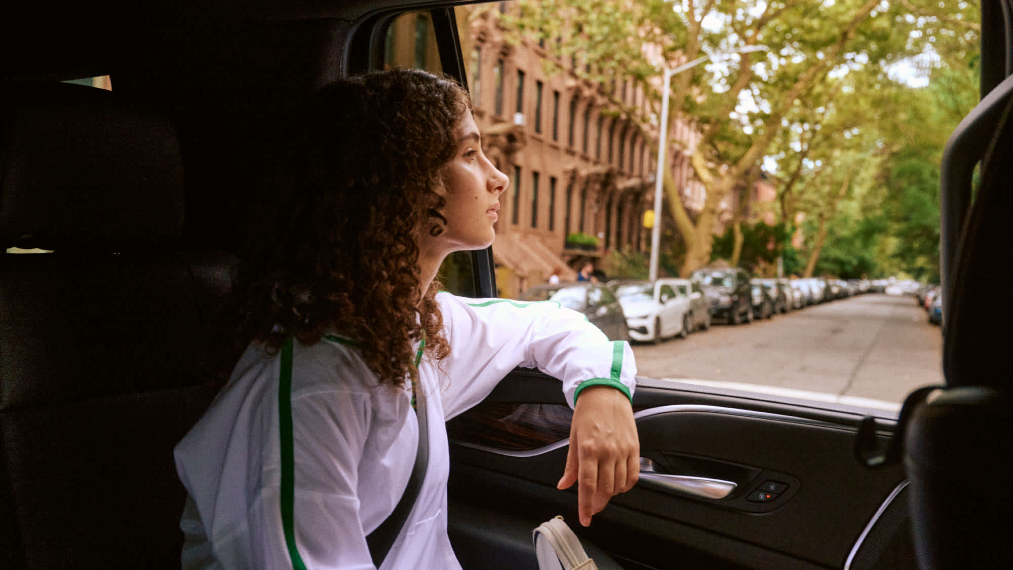 Person with curly hair in white outfit looking out car window at brownstone-lined street. 