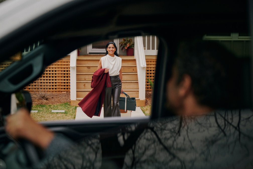 Woman in white top and maroon coat stands outside house, holding a bag, seen through a car window with driver inside.