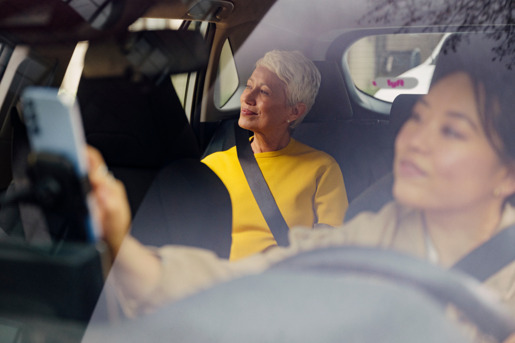 Older woman in yellow shirt sits in back seat, smiling, while driver uses a smartphone in a car with a Lyft sticker.